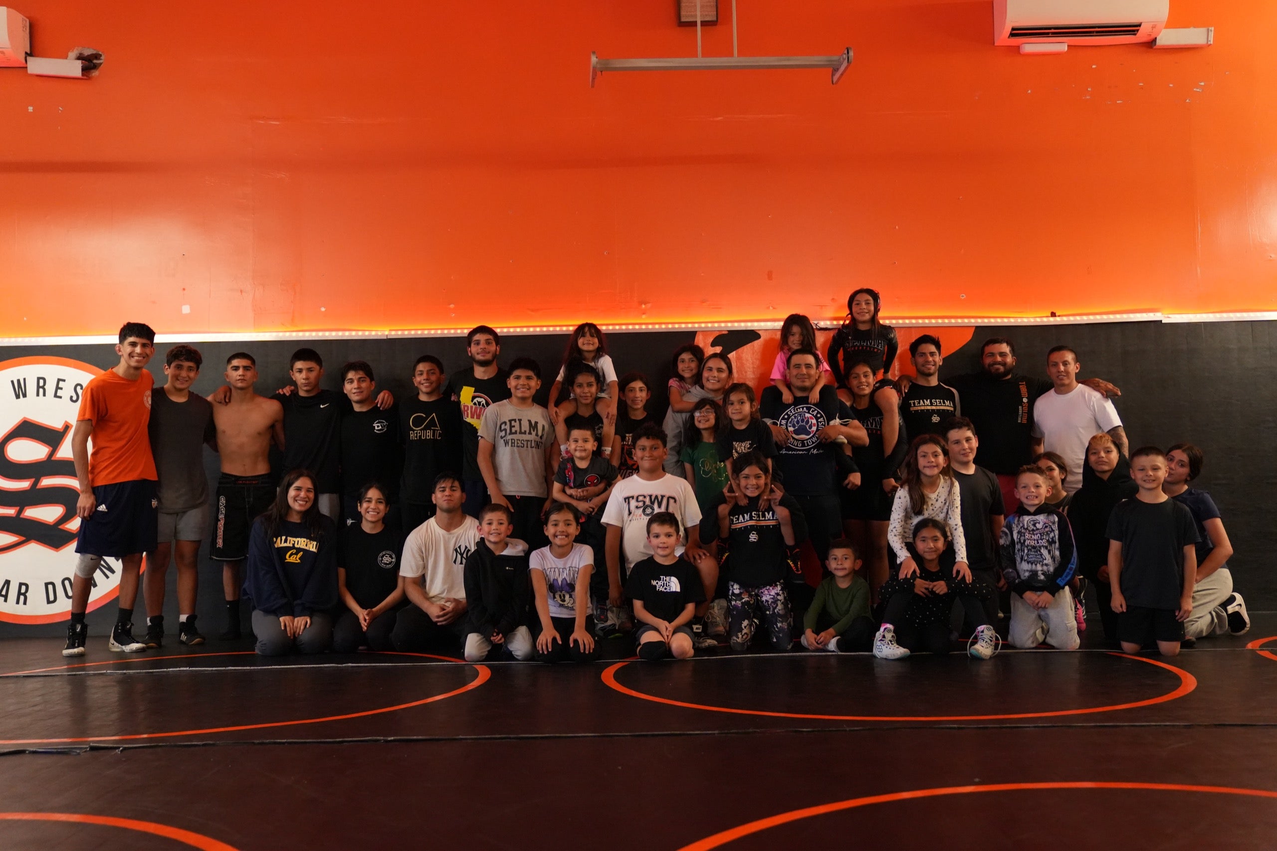 A large group of about forty-five wrestlers, children and adults, posed together in rows on an orange and black wrestling mat in a gym.