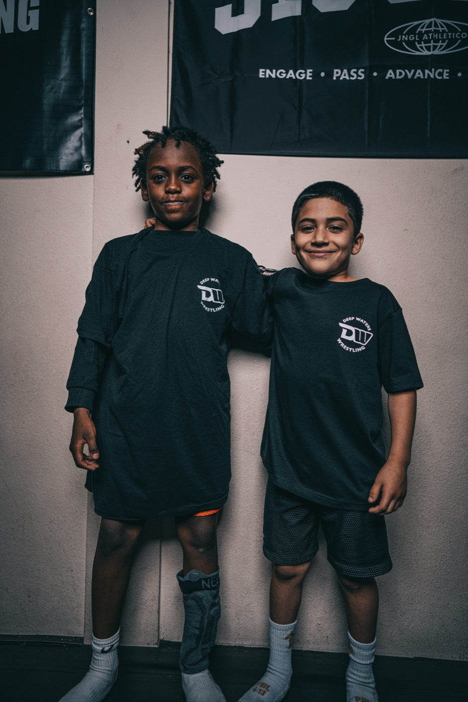 Two young boys in black club t-shirts stand side by side against a wall, one with an arm around the other, both looking at the camera and smiling.