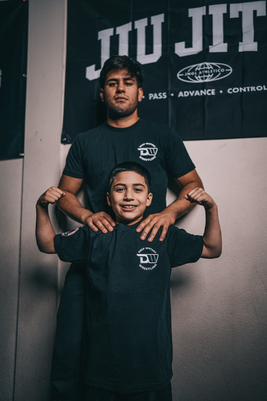 Jesse Vasquez stands against a wall in a black club t-shirt with his hands on the shoulders of a young boy in a matching oversized shirt, who flexes both arms and smiles at the camera.