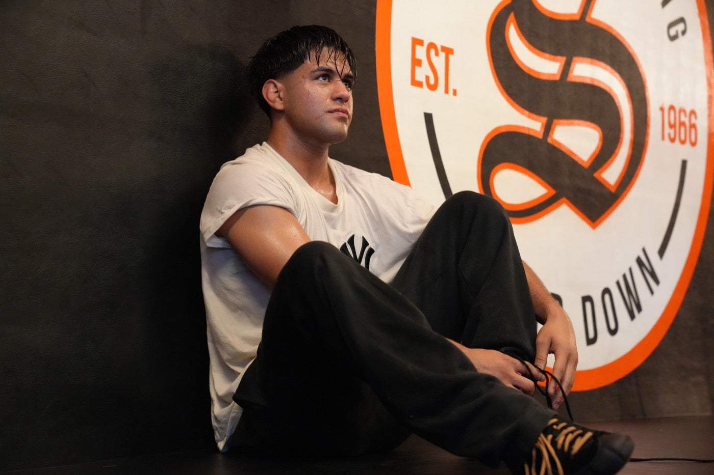 Jesse Vasquez sits on the floor against a padded wall in a sweat-soaked white t-shirt and black sweatpants, knees drawn up, tying a shoelace and looking off to his right, with a large orange gym crest on the wall behind him.