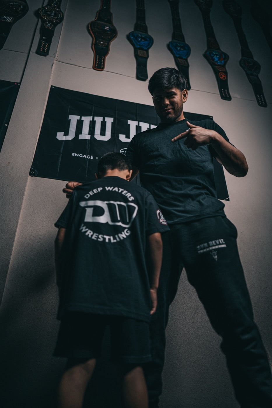 Low-angle shot of Jesse Vasquez standing against a wall hung with championship belts, pointing at the club logo on the back of a young boy's shirt; the boy faces away from the camera with his head down.