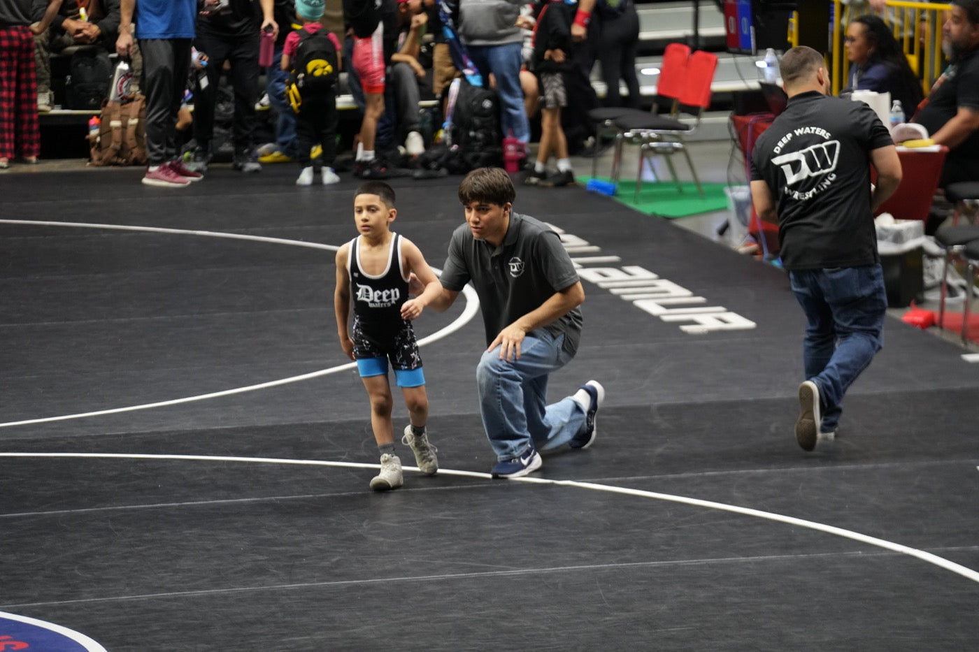 Remy Murillo kneels matside in a club polo, hand on the back of a young wrestler in a black club singlet who is walking off the mat, at an indoor tournament with spectators and a second the club coach in the background.