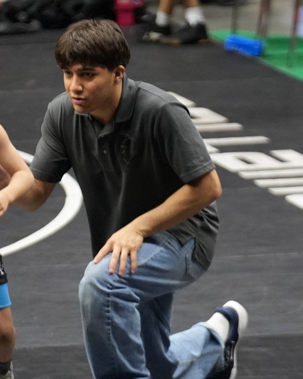 Remy Murillo kneeling at the edge of a tournament mat in a club polo, one hand reaching toward a young wrestler walking off the mat.