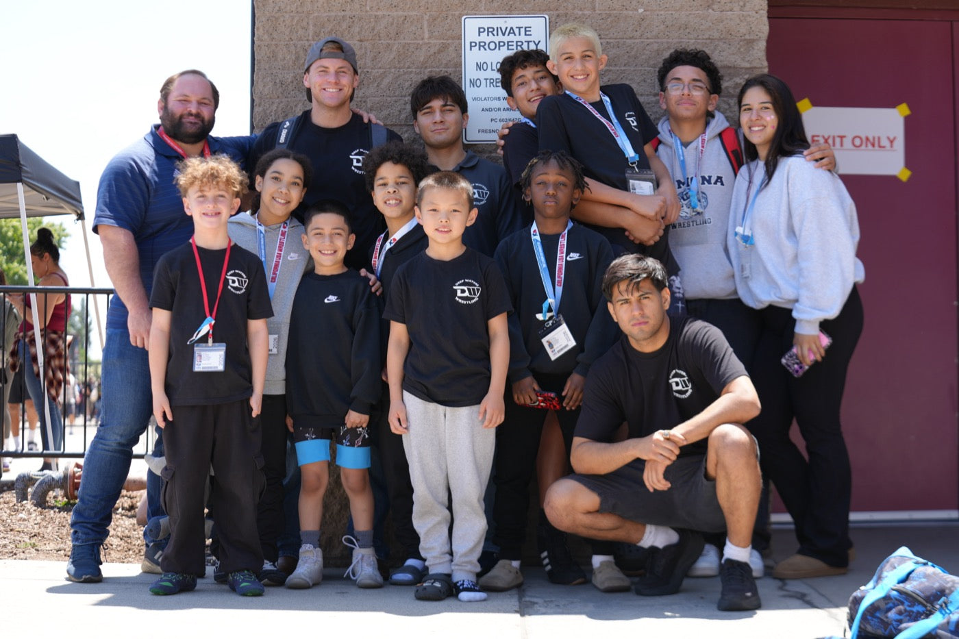 A group of about fourteen wrestlers and coaches posed together outdoors against a block wall at a tournament venue, most wearing black the club shirts, several with tournament credentials on lanyards; Jesse Vasquez crouches at the front right.