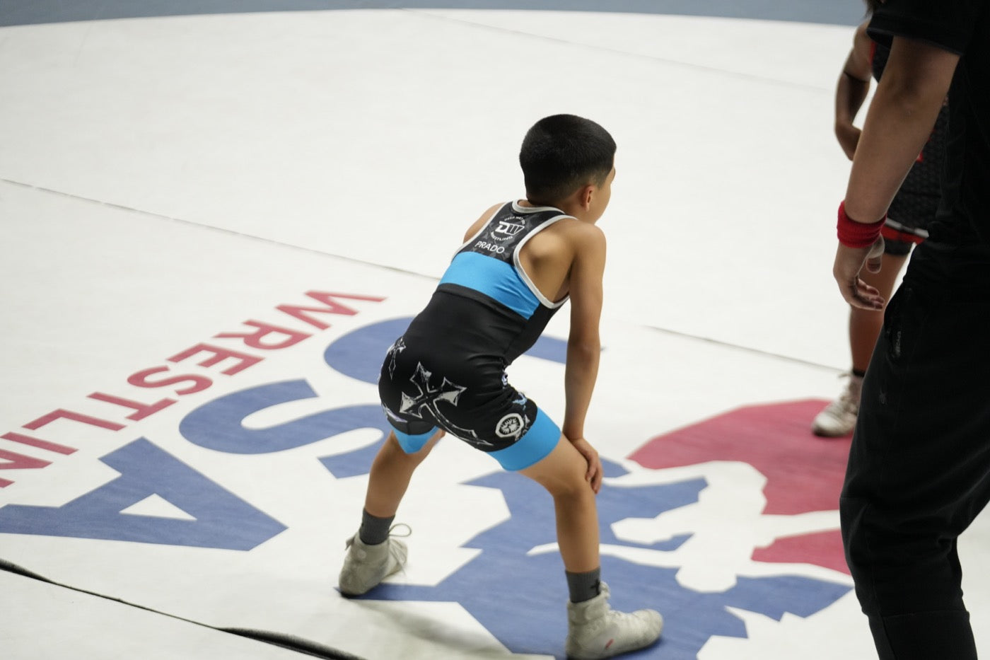 A young wrestler in a black and blue club singlet stands in a low stance on a white USA Wrestling mat, with an official and another wrestler at the edge of frame.